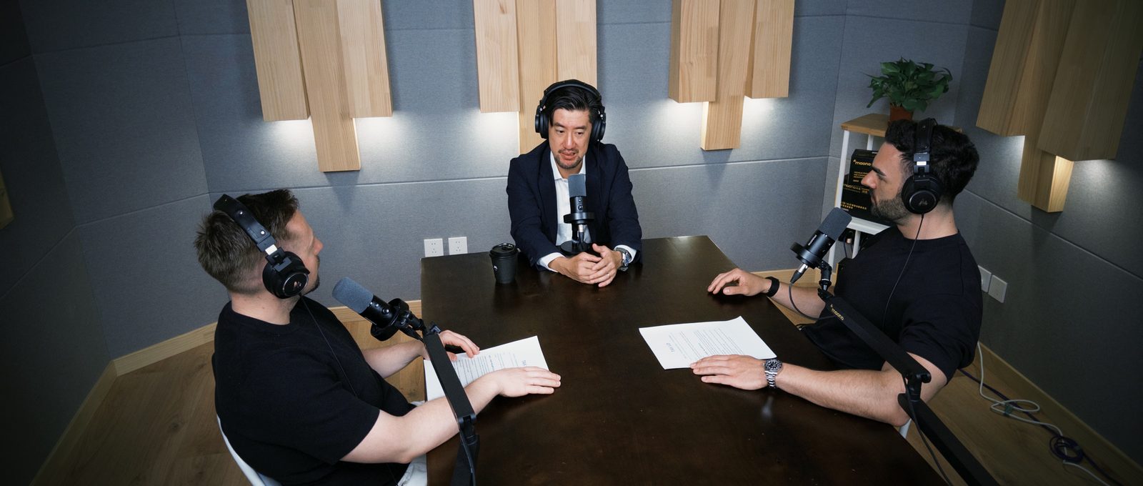 Tak Lo at the head of the table flanked by Thomas and Michael, wide three-shot opening of the Shenzhen studio recording